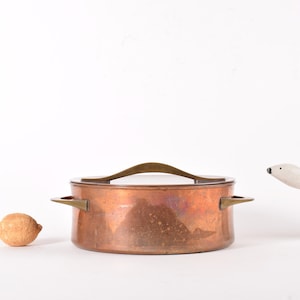 Puede incluir: Una olla de cobre con asa y tapa de latón. La olla está sobre una superficie blanca.