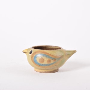 Puede incluir: Un recipiente de cerámica con forma de pájaro con un diseño amarillo, azul y marrón. El pájaro tiene un pico pequeño y un ojo redondo. El recipiente se utiliza probablemente para guardar objetos pequeños o como pieza decorativa.