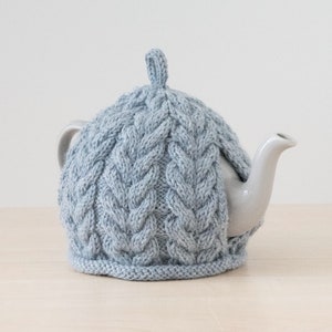 May include: A light blue knitted teapot cosy with a cable knit pattern. The cosy is on a white teapot with a spout.