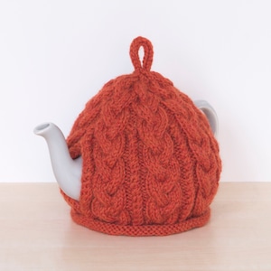 Puede incluir: Un calienta-tetera de punto naranja con un patrón de trenza. El calienta-tetera está sobre una tetera blanca con un fondo blanco.