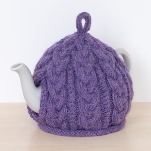 May include: A purple cable-knit tea cozy covers a gray teapot. The cozy has a loop at the top for easy handling. The teapot is on a light wood surface with a white background.