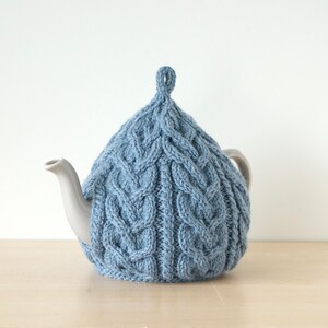 Peut inclure: Un cache-thé en tricot bleu clair avec un motif torsadé. Il recouvre une théière grise, dont le bec et l'anse sont visibles. Le cache-thé est orné d'une boucle sur le dessus pour faciliter son retrait.