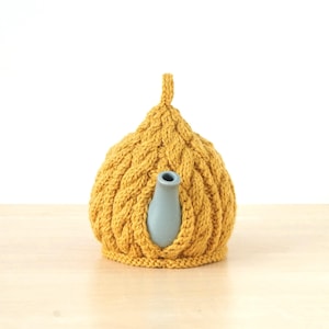 Puede incluir: Un cubrete de té tejido de color amarillo mostaza con un diseño de trenza. El cubrete cubre una tetera azul claro, con un pico y un asa visibles. El cubrete tiene un lazo en la parte superior. Fondo blanco.