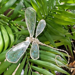 May include: A silver wire dragonfly with iridescent green wings and a pearl bead accent. The dragonfly is posed on a green leafy plant.