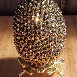 May include: A decorative egg covered in small, faceted, metallic-colored crystals. The egg sits on a gold-colored metal stand with dragonfly-shaped supports. The egg's surface reflects light, creating a sparkling effect. The overall design is ornate and elegant.