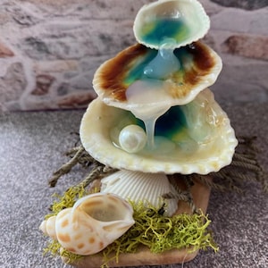 May include: A decorative seashell fountain with three stacked shells, each holding a blue and white liquid. A pearl sits in the bottom shell. The base features a seashell, moss, and rope, creating a coastal theme.