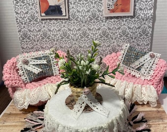 May include: A miniature living room set with two pink crocheted armchairs, a white table with a lace tablecloth, a potted plant, and a rug with black and pink fringe.