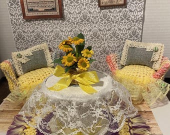 May include: A miniature living room set with two yellow crocheted armchairs, each with a gray cushion and a lace trim. A small table with a white lace tablecloth sits between the chairs. A bouquet of yellow flowers with a yellow ribbon sits on the table. The room has a gray and white patterned wallpaper with two framed pictures.