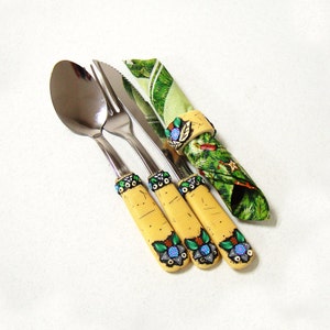 May include: Three silver utensils with yellow and blue patterned handles. The utensils are a fork, a knife, and a spoon. A green and white patterned napkin is wrapped around the handles of the utensils.