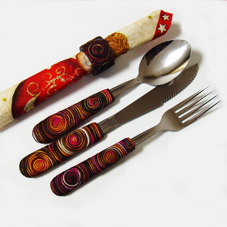 May include: A set of three silver utensils with colourful, patterned handles. The handles are made of polymer clay and feature a swirling design in shades of brown, orange, pink, and white. The utensils include a fork, a knife, and a spoon.
