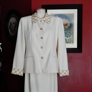 May include: Ivory-colored skirt suit with a blazer and knee-length skirt. The blazer features gold-colored buttons and trim on the collar and cuffs. The suit is displayed on a mannequin against a red wall.