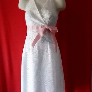 May include: White halter dress with a floral lace pattern. The dress features a pink ribbon tied around the waist. The dress is displayed on a mannequin against a red background. The dress is knee-length.