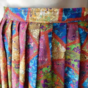 May include: A pleated skirt with a vibrant, abstract pattern. The design features a mix of colors including purple, blue, green, and gold, with red trim outlining geometric shapes. The skirt's waistband is visible at the top.