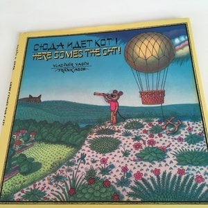 May include: A children's book titled "Here Comes The Cat!" features a colorful illustration. The cover shows a mouse with a telescope, a hot air balloon, and a cat in the background. The book has a yellow border and text in both English and Russian.