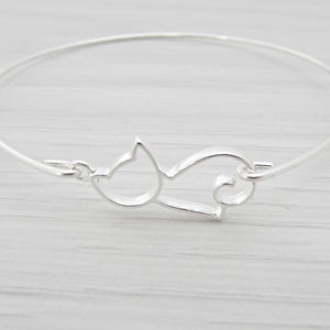 May include: A silver bangle bracelet featuring a cut-out design of a cat curled up asleep. The bracelet is simple and elegant.