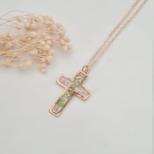 May include: A gold-colored cross pendant necklace with a delicate chain. The cross is filled with dried pink and green flowers. The necklace is displayed on a white surface, with dried flowers in the background.