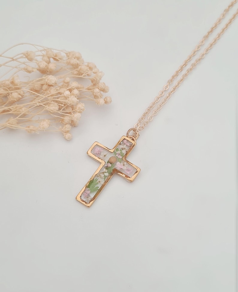 May include: A gold-colored cross pendant necklace with a delicate chain. The cross is filled with dried pink and green flowers. The necklace is displayed on a white surface, with dried flowers in the background.