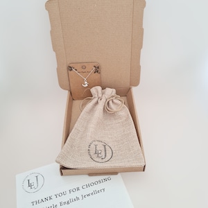 May include: A brown cardboard box with a burlap pouch inside. The pouch has a logo that says "Little English Jewellery". The box is open and a silver necklace with a small duck charm is visible. The box also includes a thank you card with the text "Thank you for choosing Little English Jewellery" and contact information.