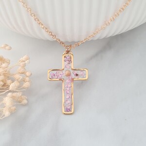 May include: A gold chain necklace with a delicate cross pendant. The cross is filled with pink and white pressed flowers.