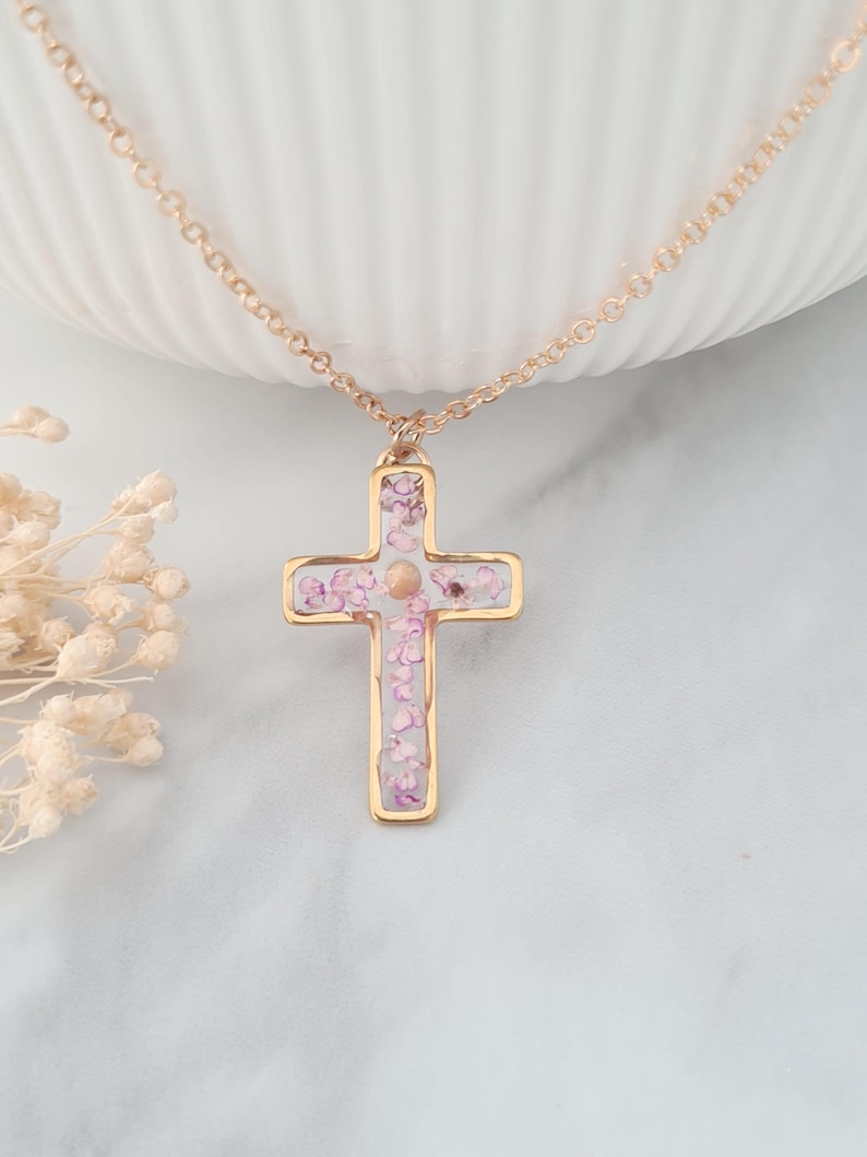 May include: A gold chain necklace with a delicate cross pendant. The cross is filled with pink and white pressed flowers.