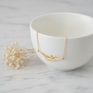 May include: A gold-coloured necklace with a bird pendant in flight. The pendant hangs from a fine chain. The necklace is displayed near a white ceramic bowl and a sprig of dried flowers.