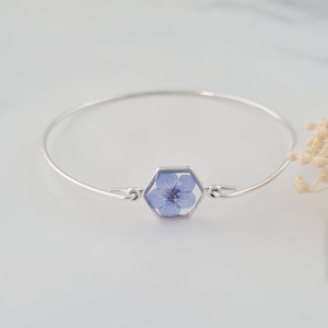 May include: A silver bangle bracelet with a hexagonal charm featuring a pressed blue flower.