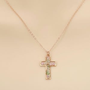 May include: A gold-colored cross pendant necklace. The cross is filled with small, dried flowers in shades of pink, purple, and green. The chain is delicate and also gold-colored. The necklace is displayed against a neutral background.