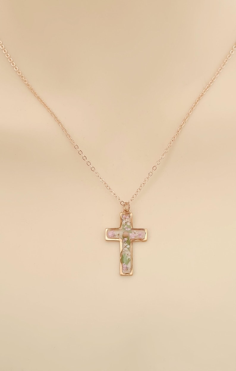 May include: A gold-colored cross pendant necklace. The cross is filled with small, dried flowers in shades of pink, purple, and green. The chain is delicate and also gold-colored. The necklace is displayed against a neutral background.