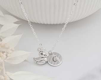 May include: A silver necklace featuring a small duck charm and a circular pendant engraved with the letter "C". The necklace is displayed on a white background, showcasing its delicate chain and charms.