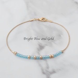 Puede incluir: Una delicada pulsera dorada con cuentas azul claro y doradas. La pulsera presenta una fina cadena y un cierre de broche. El texto "Bright Blue and Gold" se muestra en la imagen.