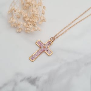 May include: A gold chain necklace with a cross pendant. The cross is filled with pink and white pressed flowers.