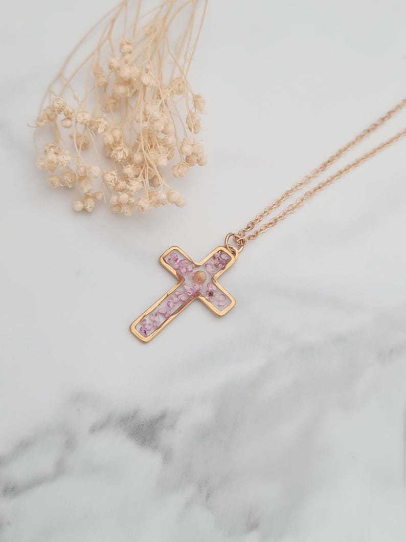 May include: A gold chain necklace with a cross pendant. The cross is filled with pink and white pressed flowers.