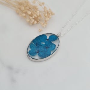 Peut inclure: Un collier argenté avec un pendentif ovale contenant des fleurs d'hortensia bleues encapsulées dans de la résine transparente. Des fleurs beiges séchées sont en arrière-plan. Le pendentif est suspendu à une chaîne délicate.