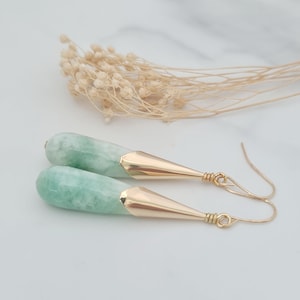 May include: A pair of drop earrings featuring smooth, pale green, cylindrical stone pendants capped with polished gold-tone metal cones. The earrings are attached to simple gold-tone wire hooks, showcasing a clean, elongated design with a subtle marbled texture on the green stone components.