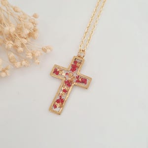 May include: Gold-tone cross pendant necklace. The cross is filled with red and gold-colored accents. The necklace has a delicate gold chain. The background is white.