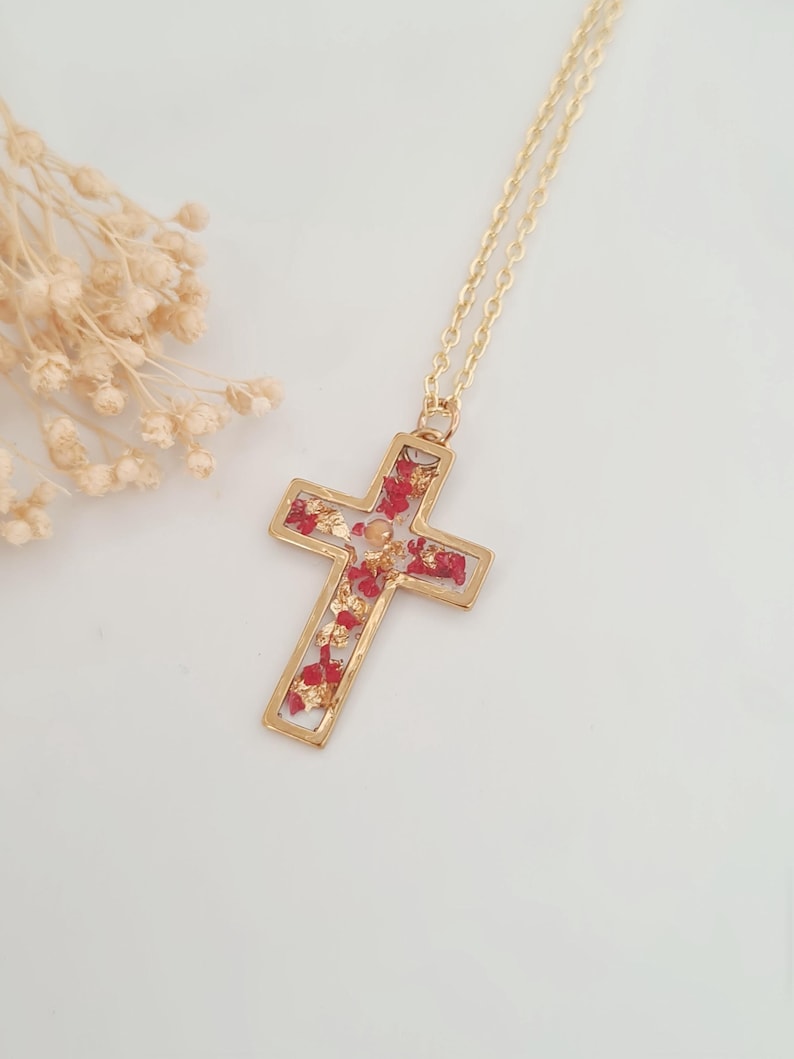May include: Gold-tone cross pendant necklace. The cross is filled with red and gold-colored accents. The necklace has a delicate gold chain. The background is white.