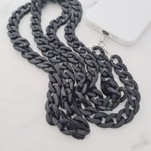 May include: A black chain phone strap with large, interlocking links. The strap is connected to a white phone case with a silver-coloured clasp. The chain is lying on a white marble surface.