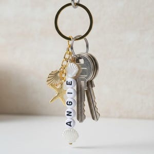 Op de afbeelding: Een goudkleurige sleutelhanger met sleutels, een gouden schelpenbedel en een zeesterbedel. De sleutelhanger heeft een streng witte kralen met de tekst "ANGIE" en schelpenbedels. De sleutelhanger hangt aan een metalen haak.