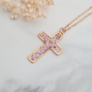 May include: A gold cross necklace with a clear resin pendant filled with pink and white dried flowers.