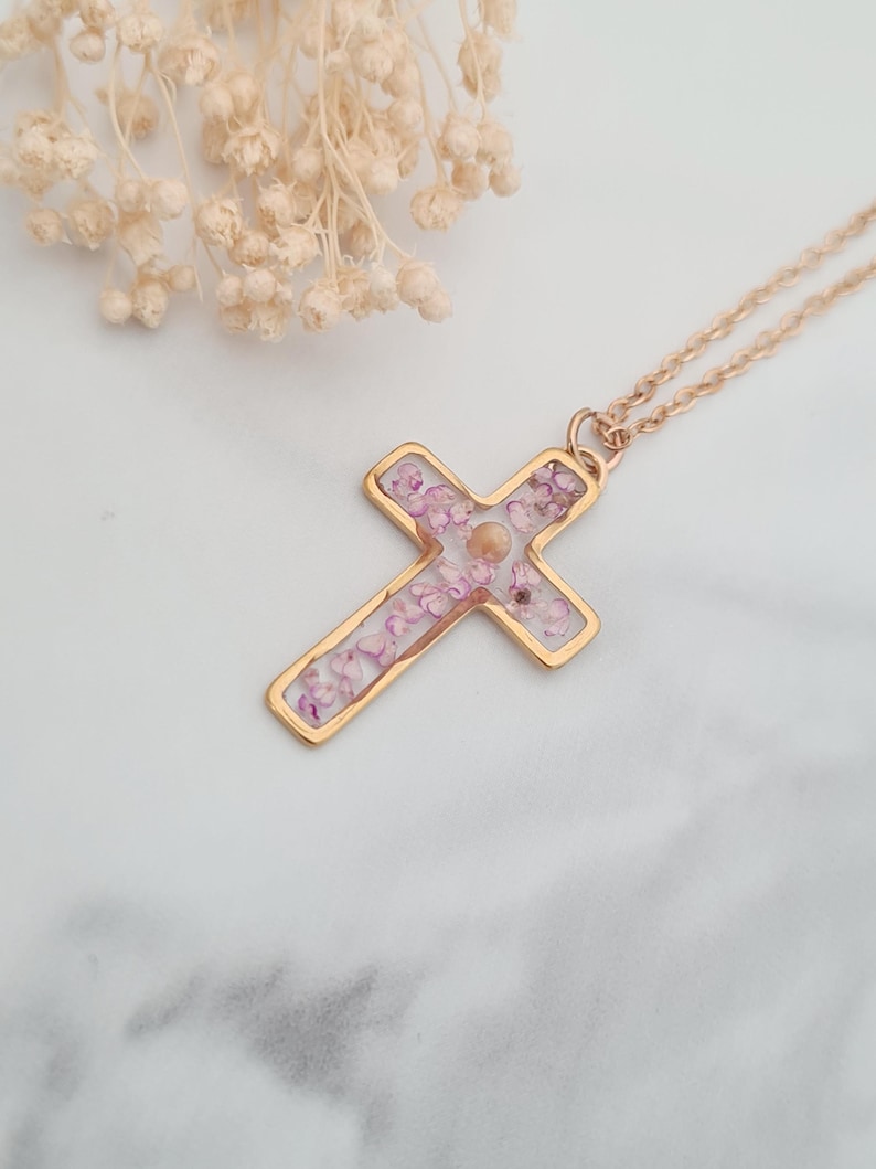 May include: A gold cross necklace with a clear resin pendant filled with pink and white dried flowers.