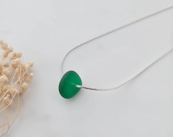 May include: A necklace featuring a single, smooth, rounded piece of vibrant green sea glass suspended on a delicate, thin silver-toned chain. The glass has a matte, frosted texture and is centered on the chain, which has a fine, metallic link design.