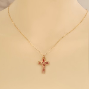 May include: A gold-colored necklace with a cross pendant. The cross is filled with small red and pink elements. The necklace is displayed on a neutral background, highlighting the jewelry's design and color.
