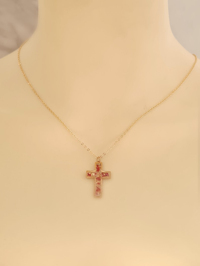 May include: A gold-colored necklace with a cross pendant. The cross is filled with small red and pink elements. The necklace is displayed on a neutral background, highlighting the jewelry's design and color.