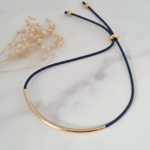 Peut inclure: Un collier composé d'une barre métallique dorée lisse et incurvée, centrée sur un cordon bleu marine foncé. Le cordon comprend un mécanisme de perle coulissante dorée réglable et deux petits embouts dorés aux extrémités du cordon.