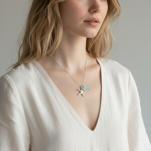 May include: A silver necklace with a starfish pendant and a light blue gemstone. The necklace is worn over a white V-neck top. The woman has blonde hair and is looking to the side.