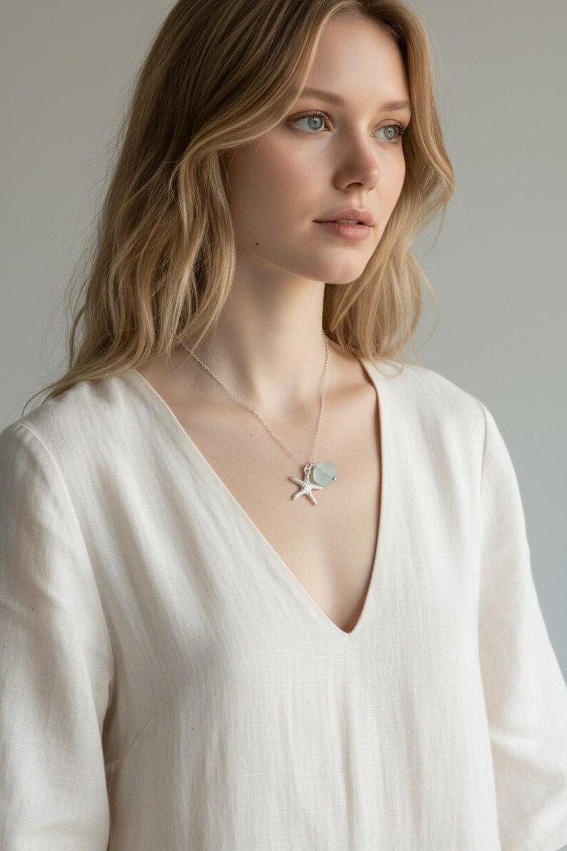 May include: A silver necklace with a starfish pendant and a light blue gemstone. The necklace is worn over a white V-neck top. The woman has blonde hair and is looking to the side.