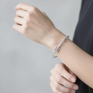 May include: A silver-coloured arrow bracelet. The arrow design has a detailed feather pattern and is worn on a wrist. The bracelet is a simple, elegant piece of jewellery.