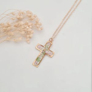 May include: A gold-colored cross pendant necklace with a delicate chain. The cross is filled with small, dried flowers in shades of pink, green, and white. The necklace is displayed on a white surface, with dried flowers in the background.