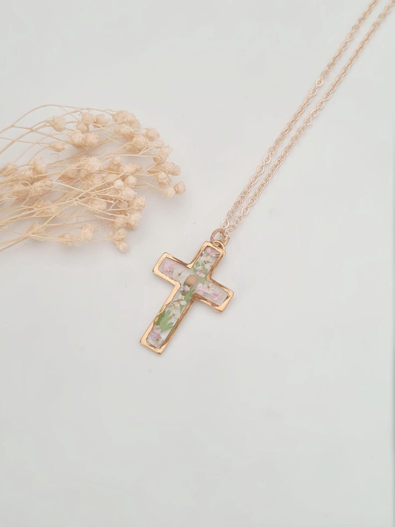 May include: A gold-colored cross pendant necklace with a delicate chain. The cross is filled with small, dried flowers in shades of pink, green, and white. The necklace is displayed on a white surface, with dried flowers in the background.