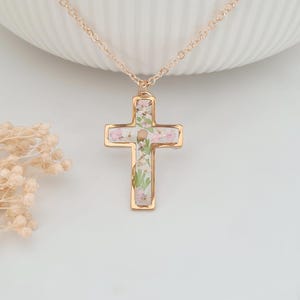 May include: A gold-colored cross pendant necklace. The cross is filled with small pink and white flowers and greenery. The necklace chain is also gold-colored. The pendant hangs against a white background.